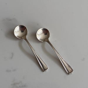 May include: Two small, silver spoons with round bowls and long, tapered handles. The spoons are placed side-by-side on a light-colored surface. The handles have a decorative, scalloped design at the base.