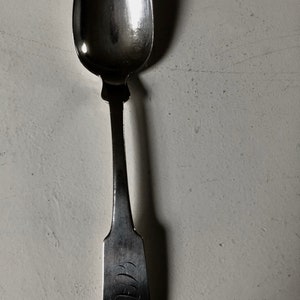 May include: A silver spoon with a rounded bowl and a long, slender handle. The handle has a decorative design and is engraved with the initials "W.B."