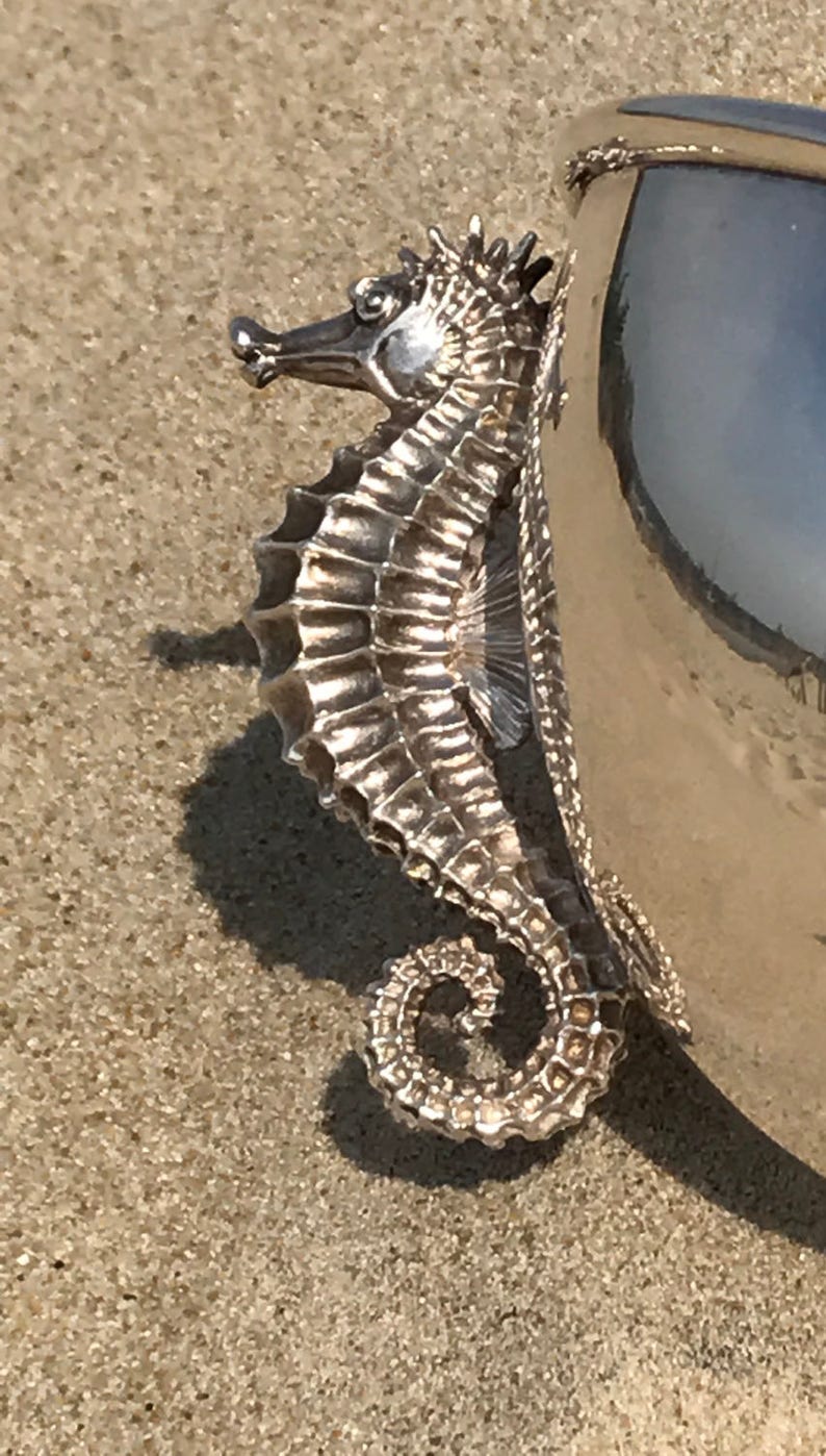 May include: Silver serving bowl with a detailed seahorse handle. The bowl is polished and reflects the surrounding environment.