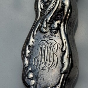 May include: Close-up of a silver-toned, ornate object with a curved shape. The object features intricate floral and foliate designs, with a central monogram of intertwined letters "MB". The metal has a slightly aged, antique appearance.