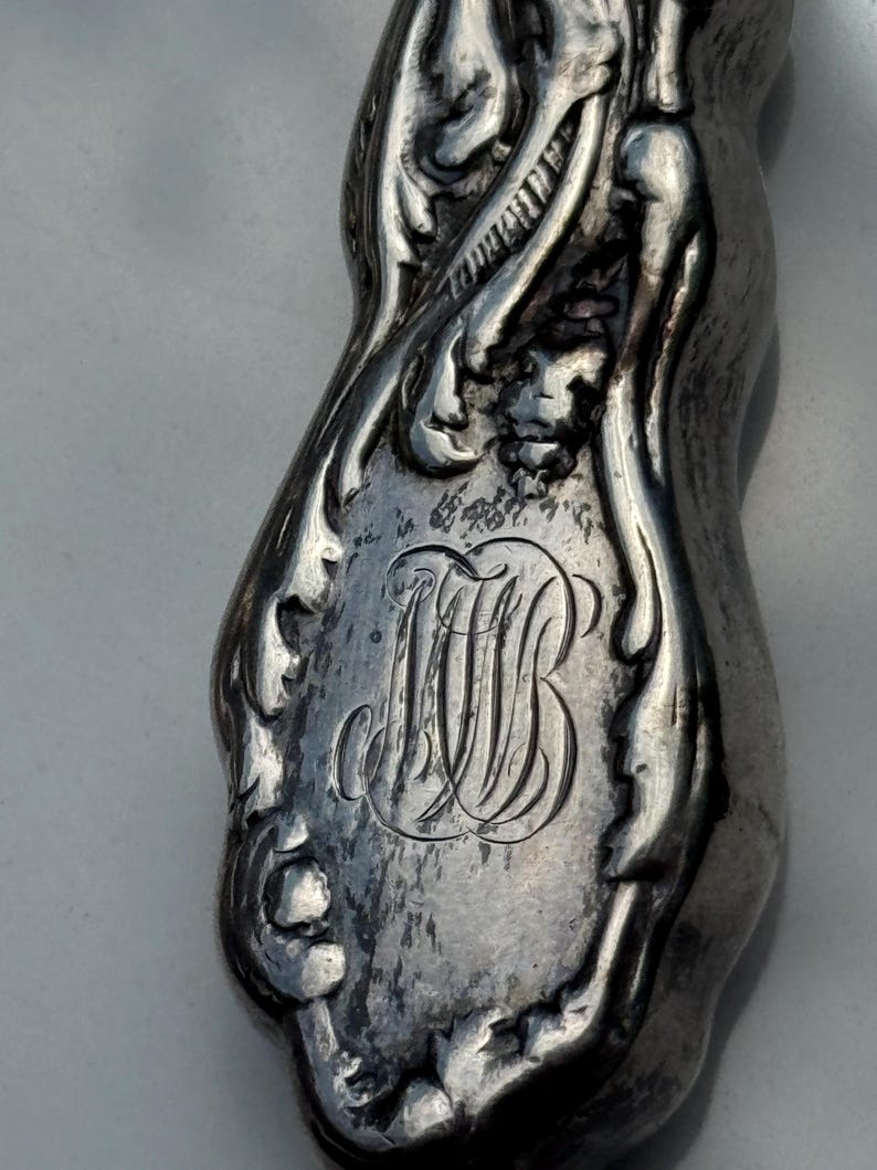 May include: Close-up of a silver-toned, ornate object with a curved shape. The object features intricate floral and foliate designs, with a central monogram of intertwined letters "MB". The metal has a slightly aged, antique appearance.