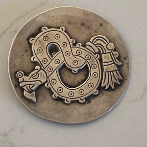 May include: A silver circular pendant with an embossed design of a serpent with a feathered headdress. The serpent is curled into a circle with its tail in its mouth.