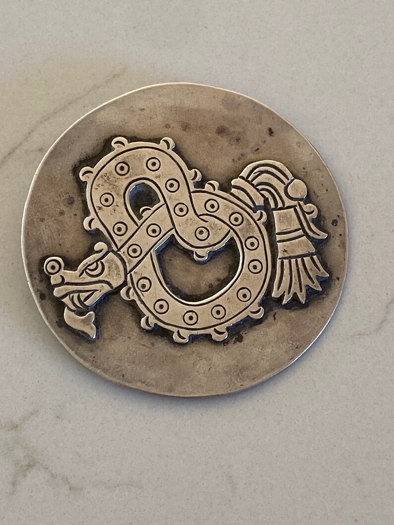 May include: A silver circular pendant with an embossed design of a serpent with a feathered headdress. The serpent is curled into a circle with its tail in its mouth.