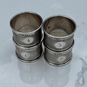 May include: Four silver napkin rings stacked in two pairs. Each ring has a textured band and a small, numbered oval. The numbers 1, 2, 3, and 4 are visible. The rings are on a reflective surface.