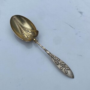 May include: A silver spoon with an ornate design. The bowl of the spoon is oval-shaped and has a floral pattern. The handle is decorated with a vine and berries.