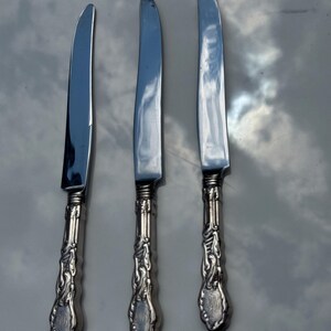 May include: Three silver knives with decorative handles. The blades are smooth and reflective, and the handles have a detailed, scrolling design with a monogrammed oval at the base. The knives are arranged in a row.