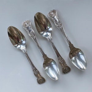 Puede incluir: Cuatro cucharas de plata con mangos ornamentados. Las cucharas están dispuestas en fila, con los mangos apuntando hacia la parte superior de la imagen.