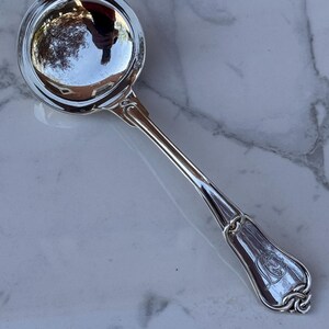 May include: A shiny, silver-colored ladle with a round bowl and a long, slender handle. The handle features decorative detailing and a monogram at the end. The ladle is resting on a white and gray marble surface.