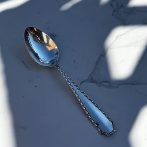 Puede incluir: Una cuchara plateada con una superficie reflectante, apoyada sobre una superficie de mármol blanco y gris. El mango de la cuchara tiene un diseño decorativo. La cuchara está orientada hacia la esquina superior izquierda de la imagen.