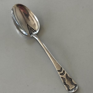 Peut inclure: Une cuillère en argent avec un bol ovale et un long manche fin. La poignée est ornée de motifs gravés près de l'extrémité. La cuillère est posée sur un fond gris.