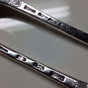 May include: Close-up of two antique silver spoons. The spoons have ornate, engraved floral designs on the handles. The silver spoons have several hallmarks and maker's marks stamped into the metal. The spoons are on a white surface.