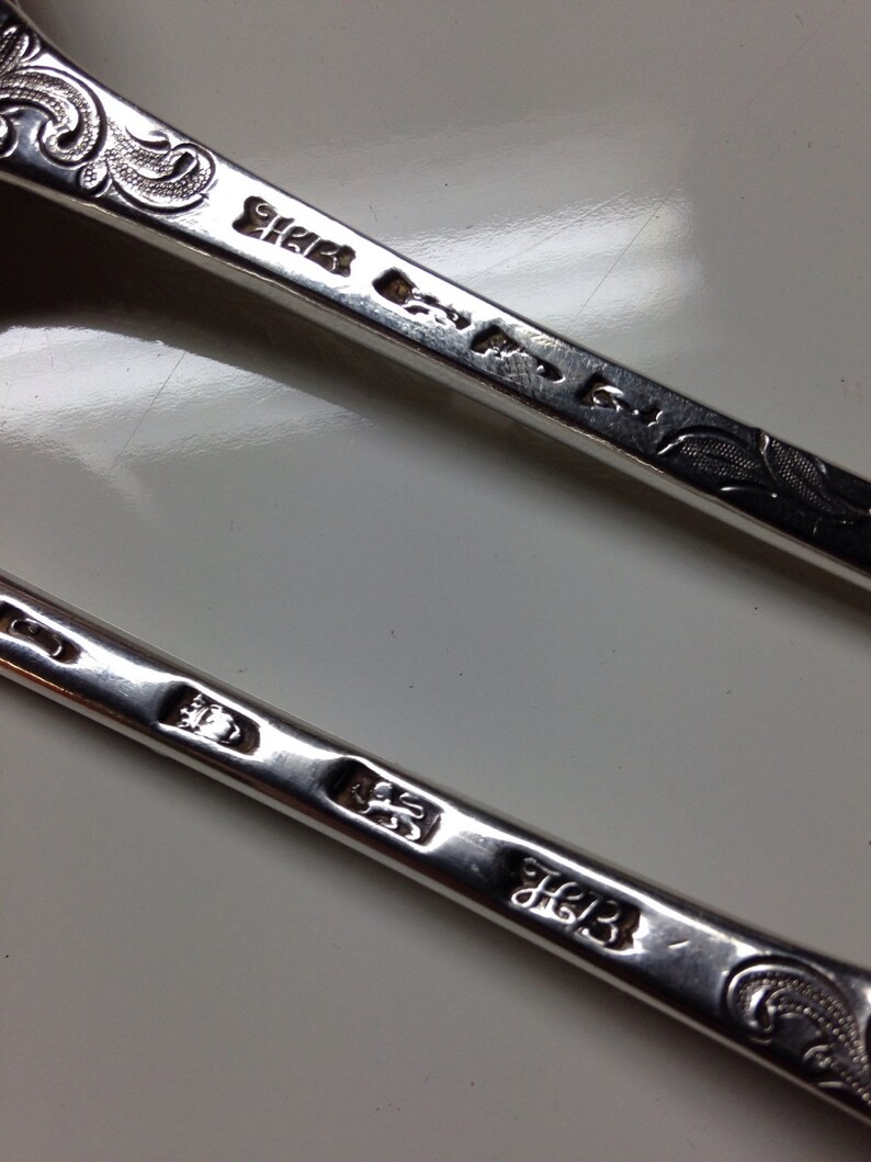 May include: Close-up of two antique silver spoons. The spoons have ornate, engraved floral designs on the handles. The silver spoons have several hallmarks and maker's marks stamped into the metal. The spoons are on a white surface.