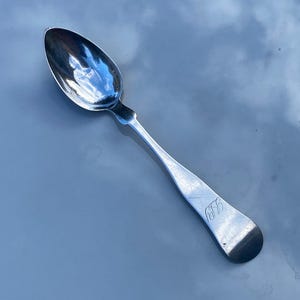 Puede incluir: Una cuchara de plata con un cuenco redondeado y un mango delgado. El mango tiene un elemento decorativo cerca del cuenco y un monograma en el extremo. La cuchara es reflectante.