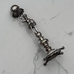Peut inclure: Un chandelier en argent avec une figurine d'une personne tenant un petit objet au sommet. Le chandelier a une base ronde avec trois pieds.