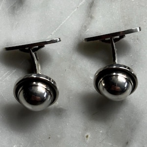 May include: A pair of silver cufflinks with a spherical design. Each cufflink features a polished, rounded ball shape encased in a circular frame, connected to a rectangular bar. The cufflinks are displayed on a light gray, marbled surface.