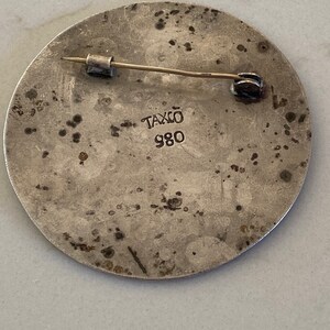 May include: Silver circular pin with a textured surface and a pin back. The pin has the text "Taxco 980" on the back.