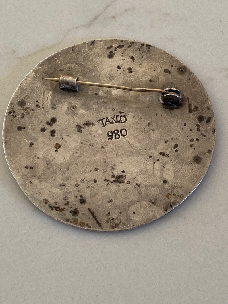 May include: Silver circular pin with a textured surface and a pin back. The pin has the text "Taxco 980" on the back.