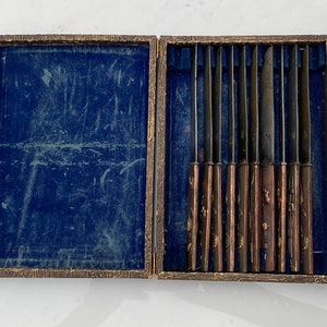 K&ouml;nnte beinhalten: Eine Holzkiste mit blauer Samtf&uuml;tterung enth&auml;lt ein Set von acht silbernen Messern mit verzierten Griffen.
