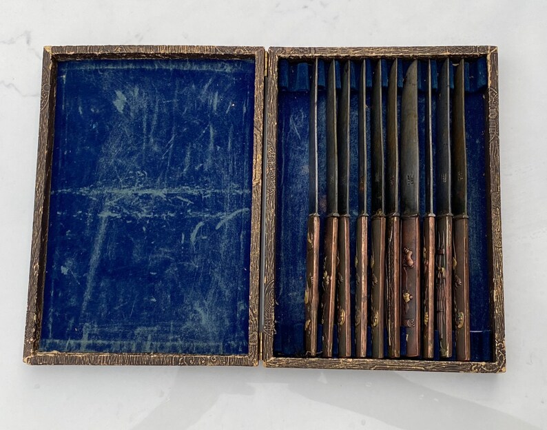 K&ouml;nnte beinhalten: Eine Holzkiste mit blauer Samtf&uuml;tterung enth&auml;lt ein Set von acht silbernen Messern mit verzierten Griffen.