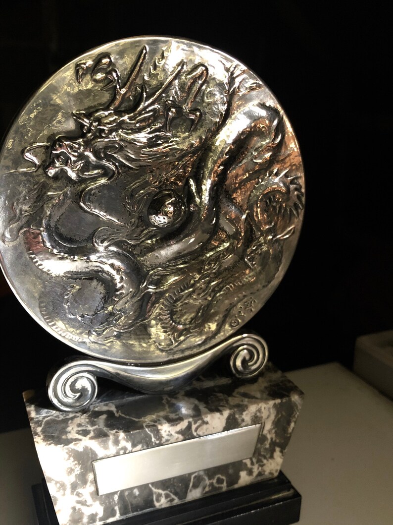 May include: A silver trophy with a dragon design on a round base. The base is attached to a marble base with a silver plaque.