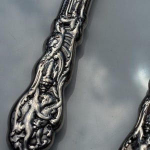 May include: Close-up of ornate silver cutlery. The handle features detailed relief carvings of figures and floral patterns. The silver has a polished finish, reflecting light. The design is intricate and classic, suggesting antique silverware.