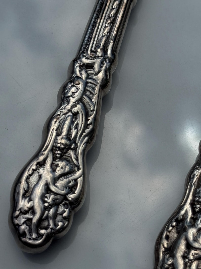 May include: Close-up of ornate silver cutlery. The handle features detailed relief carvings of figures and floral patterns. The silver has a polished finish, reflecting light. The design is intricate and classic, suggesting antique silverware.