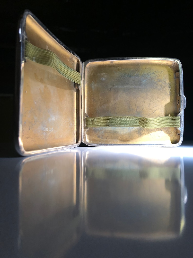 Pu&ograve; includere: Un porta sigarette in argento aperto con un elastico verde all'interno. Il porta sigarette ha un aspetto usurato e invecchiato.
