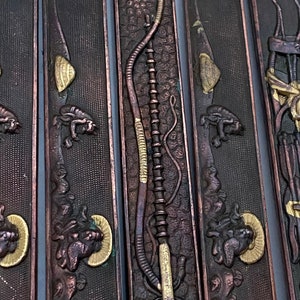 K&ouml;nnte beinhalten: Nahaufnahme eines Satzes von f&uuml;nf dekorativen Metallplatten mit aufwendigen Designs. Die Platten sind dunkelbraun mit goldenen Akzenten und zeigen eine Vielzahl von Motiven, darunter eine Schlange, Pfeil und Bogen und ein stilisiertes Tier.