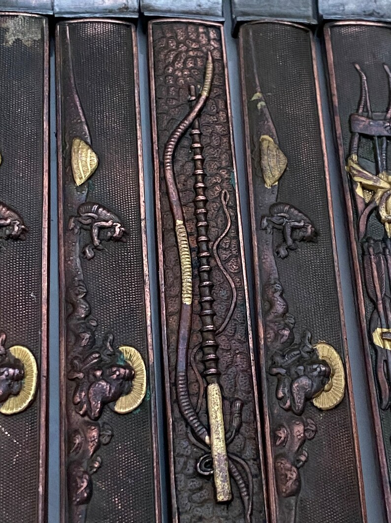 K&ouml;nnte beinhalten: Nahaufnahme eines Satzes von f&uuml;nf dekorativen Metallplatten mit aufwendigen Designs. Die Platten sind dunkelbraun mit goldenen Akzenten und zeigen eine Vielzahl von Motiven, darunter eine Schlange, Pfeil und Bogen und ein stilisiertes Tier.