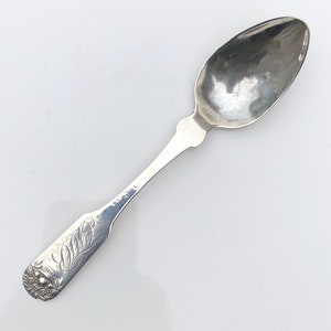 May include: A silver teaspoon with an ornate handle. The handle is decorated with a floral design and a monogram.