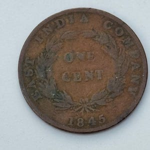 May include: A copper-colored East India Company one-cent coin from 1845. The coin features the text "EAST INDIA COMPANY" around the top edge, with "ONE CENT" in the center, surrounded by a wreath.