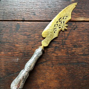 May include: A silver and gold butter knife with an ornate design. The handle is silver and the blade is gold with a cut-out design.