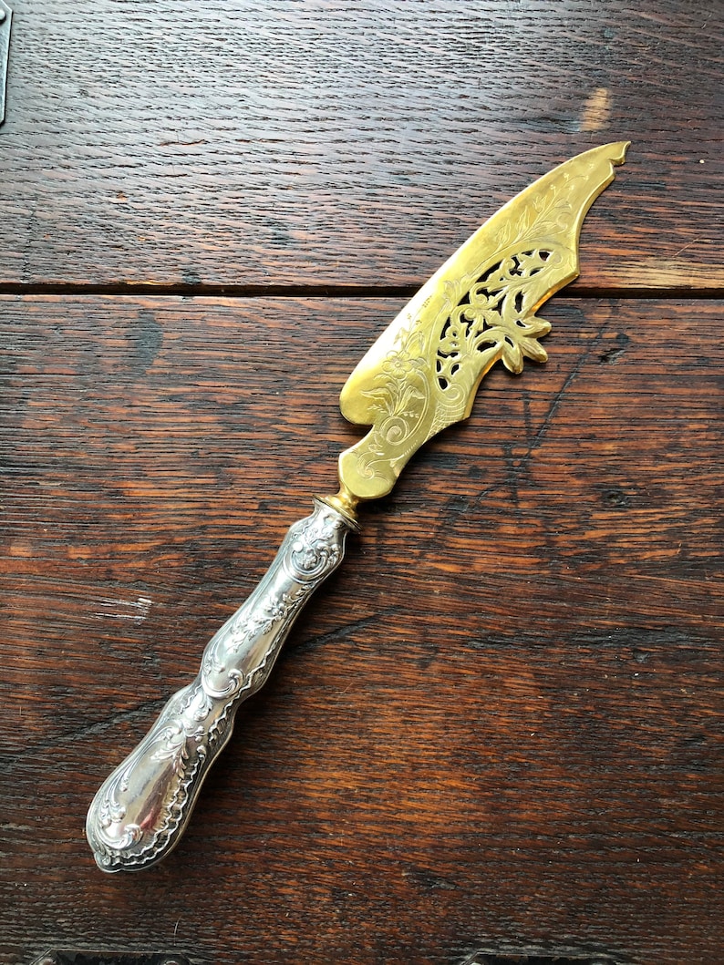 May include: A silver and gold butter knife with an ornate design. The handle is silver and the blade is gold with a cut-out design.
