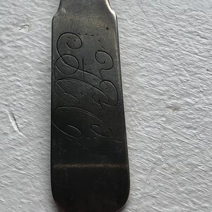 Puede incluir: Primer plano de la parte posterior de una cuchara de plata con un dise&ntilde;o grabado. El dise&ntilde;o es una serie de remolinos y bucles.