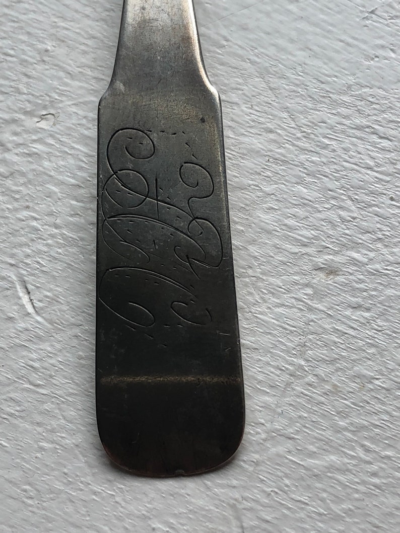 Puede incluir: Primer plano de la parte posterior de una cuchara de plata con un dise&ntilde;o grabado. El dise&ntilde;o es una serie de remolinos y bucles.