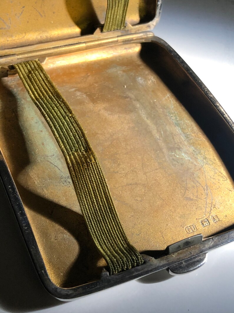 Pu&ograve; includere: Un porta sigarette d'argento con un elastico verde all'interno. La custodia ha un aspetto usurato e invecchiato e l'iscrizione "CU 21 A" &egrave; visibile sul fondo.