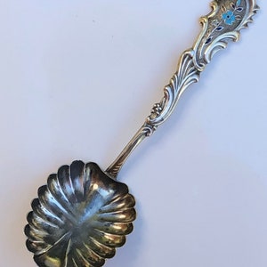 May include: A silver sugar spoon with a scalloped bowl and a decorative handle with a blue and white enamel flower design.
