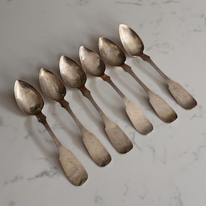 May include: Six antique silver spoons. Each spoon features an oval bowl and a slender handle, with engraved initials. The spoons are arranged diagonally on a light surface. The spoons are likely made of silver.