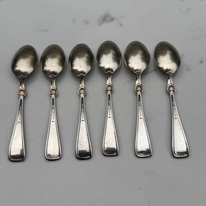 May include: Six silver spoons with oval bowls and slender handles. Each spoon has a decorative element where the bowl meets the handle and a small inscription on the handle. The spoons are arranged in a row on a light-colored surface.
