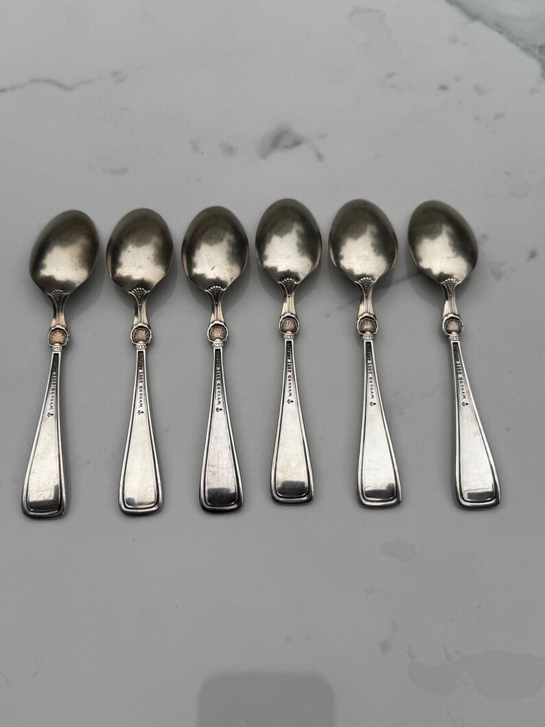 May include: Six silver spoons with oval bowls and slender handles. Each spoon has a decorative element where the bowl meets the handle and a small inscription on the handle. The spoons are arranged in a row on a light-colored surface.