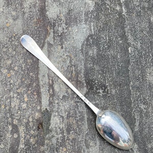 May include: A silver teaspoon with a long, slender handle and a rounded bowl. The spoon is lying on a gray, textured surface.