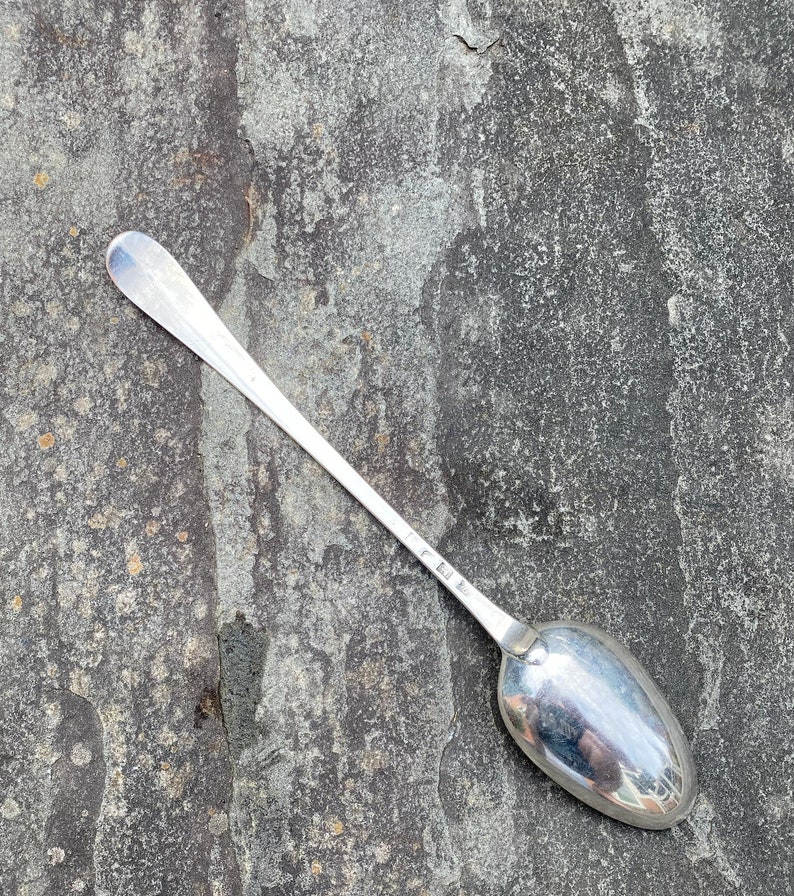 May include: A silver teaspoon with a long, slender handle and a rounded bowl. The spoon is lying on a gray, textured surface.