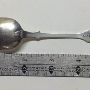 Puede incluir: Una cucharita de plata con un cuenco redondeado y un mango largo y delgado. El mango tiene un dise&ntilde;o decorativo en el extremo y la cuchara est&aacute; marcada con las letras "SOLUD". La cuchara se mide con una regla, mostrando que tiene aproximadamente 12,7 cm de largo.