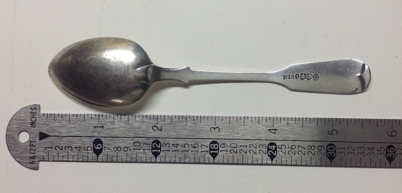 Puede incluir: Una cucharita de plata con un cuenco redondeado y un mango largo y delgado. El mango tiene un dise&ntilde;o decorativo en el extremo y la cuchara est&aacute; marcada con las letras "SOLUD". La cuchara se mide con una regla, mostrando que tiene aproximadamente 12,7 cm de largo.