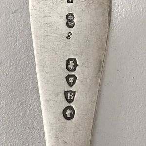 Może przedstawiać: Zbliżenie srebrnej łyżki z zaokrągloną czarką. Powierzchnia łyżki jest odblaskowa, a na trzonku wybito kilka cech probierczych, w tym 'V' w prostokącie, 'GA', '8' i inne symbole.