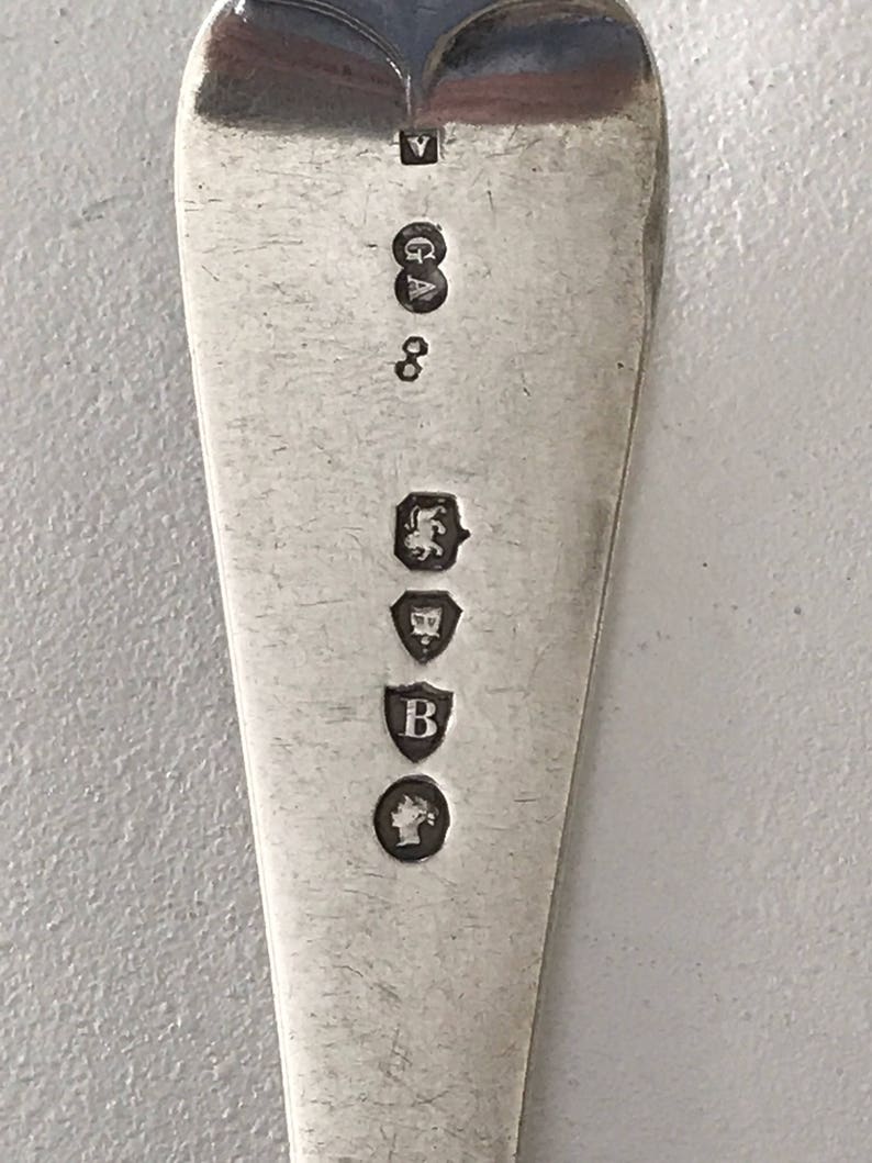 Może przedstawiać: Zbliżenie srebrnej łyżki z zaokrągloną czarką. Powierzchnia łyżki jest odblaskowa, a na trzonku wybito kilka cech probierczych, w tym 'V' w prostokącie, 'GA', '8' i inne symbole.