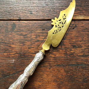 May include: A silver-handled butter knife with a brass blade featuring a decorative cutout design. The blade is ornate and has a floral pattern.