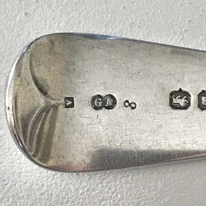 Może przedstawiać: Zbliżenie srebrnej łyżki ze znakami probierczymi. Łyżka ma zaokrągloną miseczkę i gładką, odblaskową powierzchnię. Widoczne znaki probiercze to kwadratowy znak, litery "GA" i symbol nieskończoności. Łyżka to prawdopodobnie antyczne srebro.