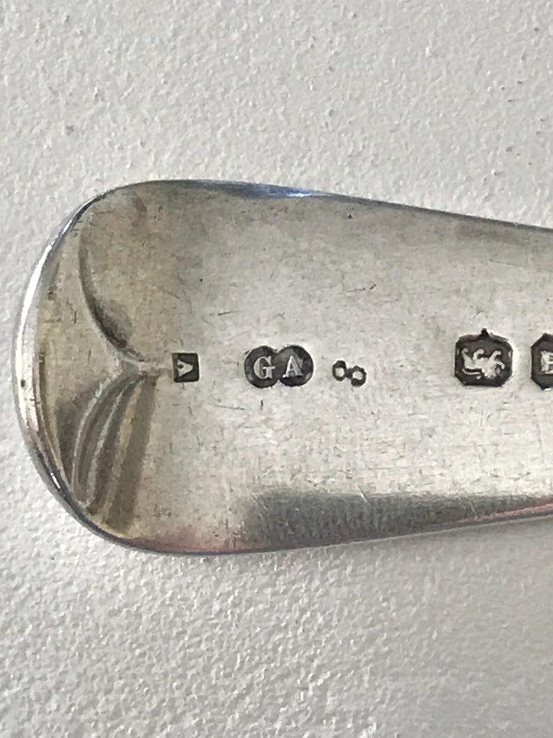 Może przedstawiać: Zbliżenie srebrnej łyżki ze znakami probierczymi. Łyżka ma zaokrągloną miseczkę i gładką, odblaskową powierzchnię. Widoczne znaki probiercze to kwadratowy znak, litery "GA" i symbol nieskończoności. Łyżka to prawdopodobnie antyczne srebro.
