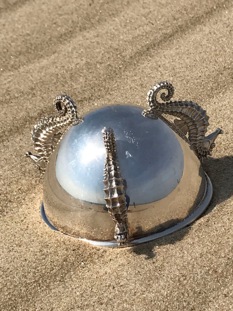 May include: A silver bowl with a shiny surface and three seahorse handles. The bowl is sitting on a sandy surface.